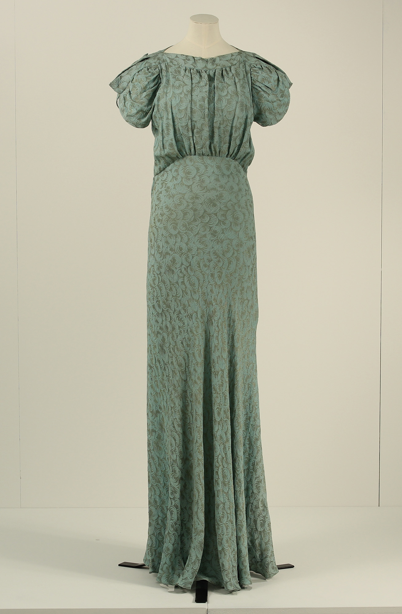 A full length, patterned green dress from the 1930s, with a fitted waist and puffed sleeves