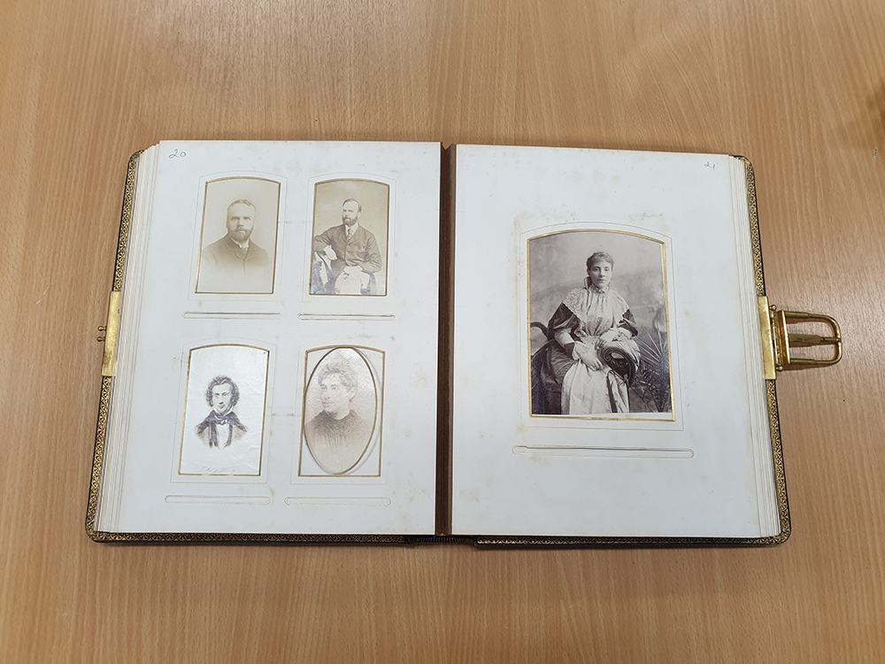 An open photo album with gilt edging and a metal buckle fastening. On the left hand page are Edwardian sepia portrait photographs of four men, the top two of whom have beards. On the right hand side is a larger photo of a young woman in a heavy, long-sleeved dress with a white lace trim around the collar and cuffs.