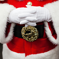 A close up of Santa's white gloved hands folded across his tummy. He's dressed in his usual fur-trimmed red coat with a black belt featuring a reindeer buckle