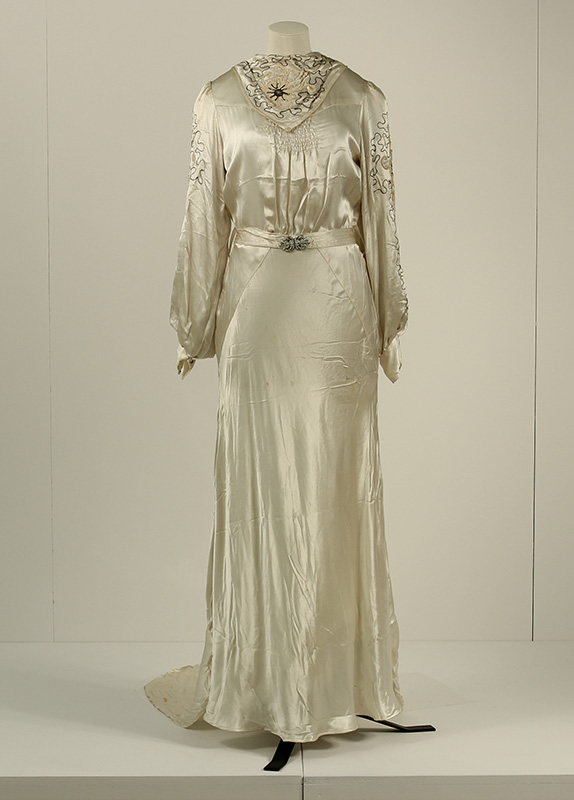 A 1930s ivory satin wedding dress with a full length skirt, long, puffed sleeves, embroidery around the neck and sleeves and a fitted waist with a decorative belt buckle