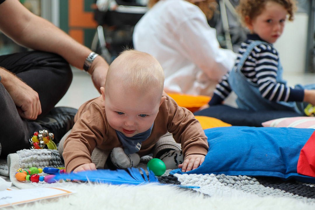 A baby sitting on a rug on the ground in the Herbert's covered court, playing with a blue feather, green rubber egg and a set of coloured beads