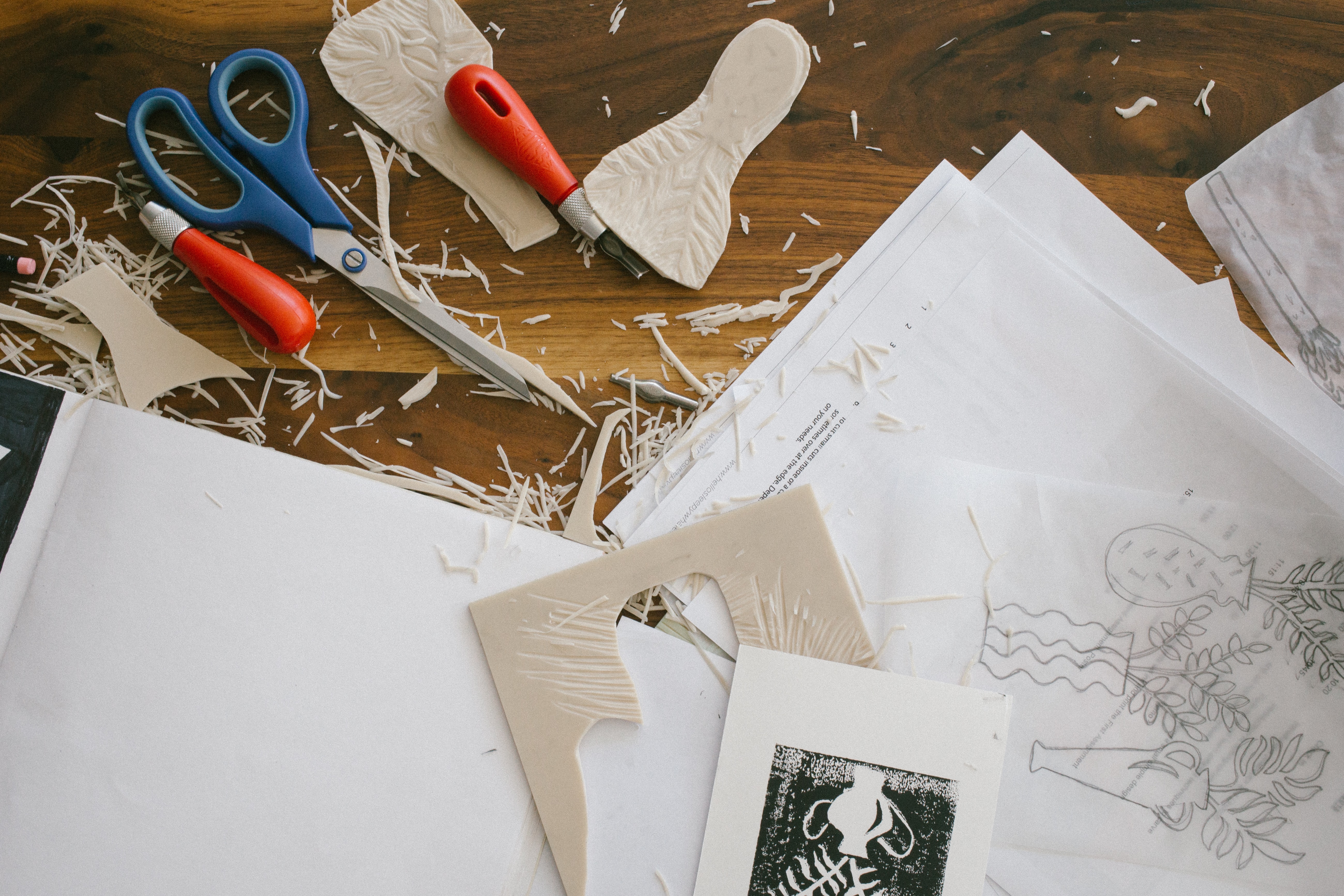 A wooden table with a linocut stamp and print, and pieces of cut out lino scattered around. There are sheets of paper, a pen, a pair of scissors and other utensils, as well as a tracing paper sketch.