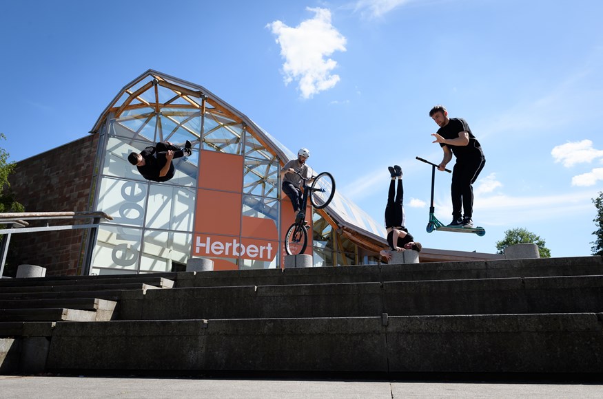 A photo of stunt performers on the steps of the Herbert Art Gallery as part of Coventry moves. On the left hand side, one performs a flip in midair. Next to him, a BMX biker balances on the back wheel of his bike on top of a bollard. Towards the right, a performer rests his head against a bollard, pushing his legs up in the air, and on the far right, the final performer is captured on a scooter in mid-air. 