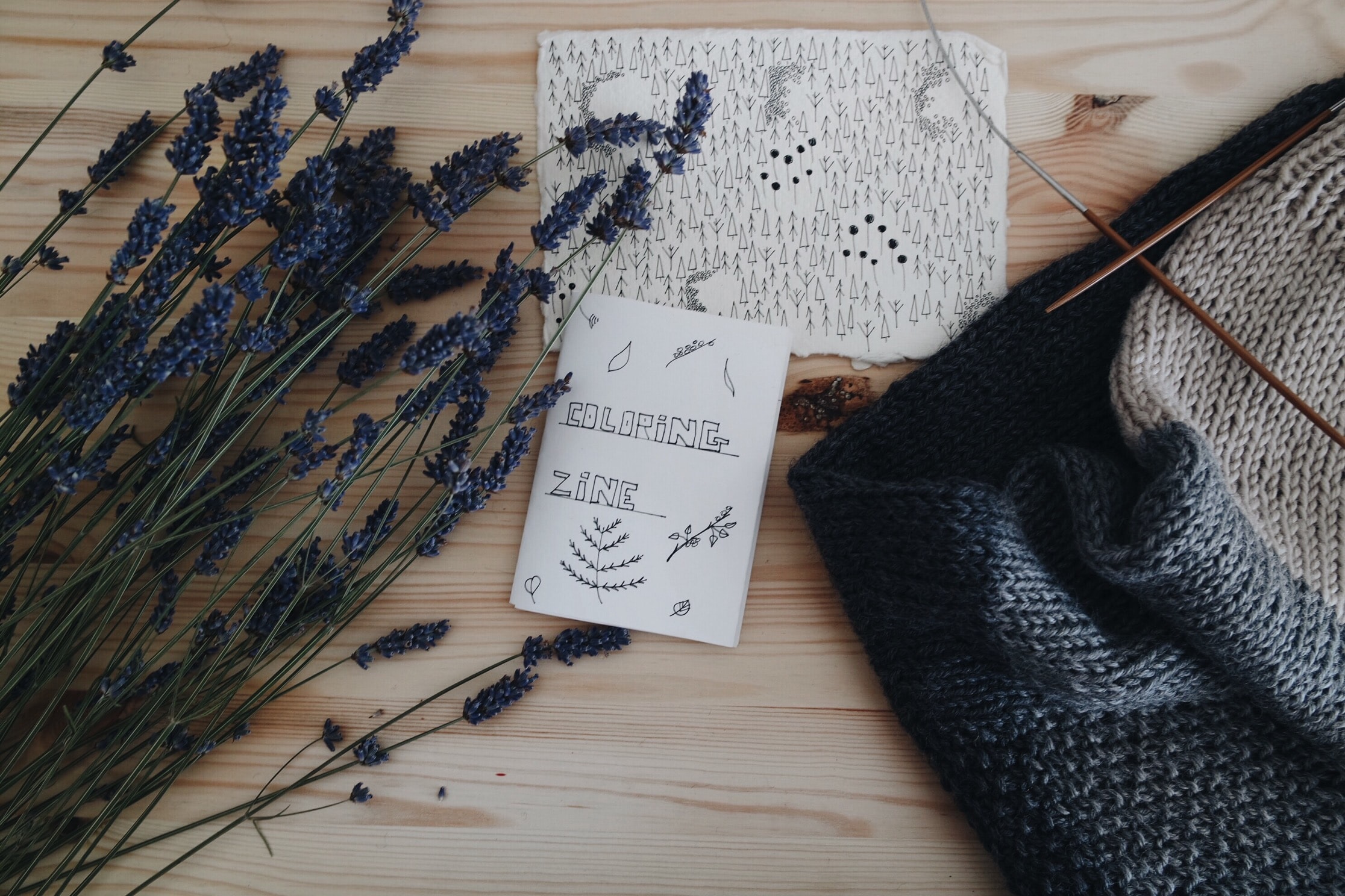 A small, hand-drawn paper zine is laid out on a wooden table next to some stalks of lavender and a wool scarf