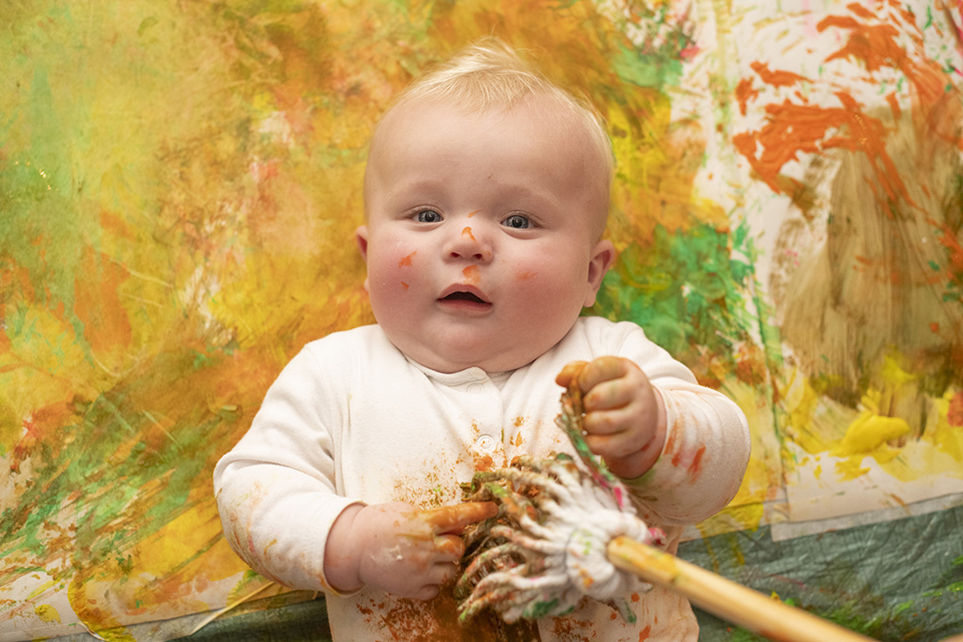 A baby lying on a mat covered with paint, with splodges of orange and yellow paint on its face, hands and tummy. Its tummy is being tickled with a mini mop covered in paint.