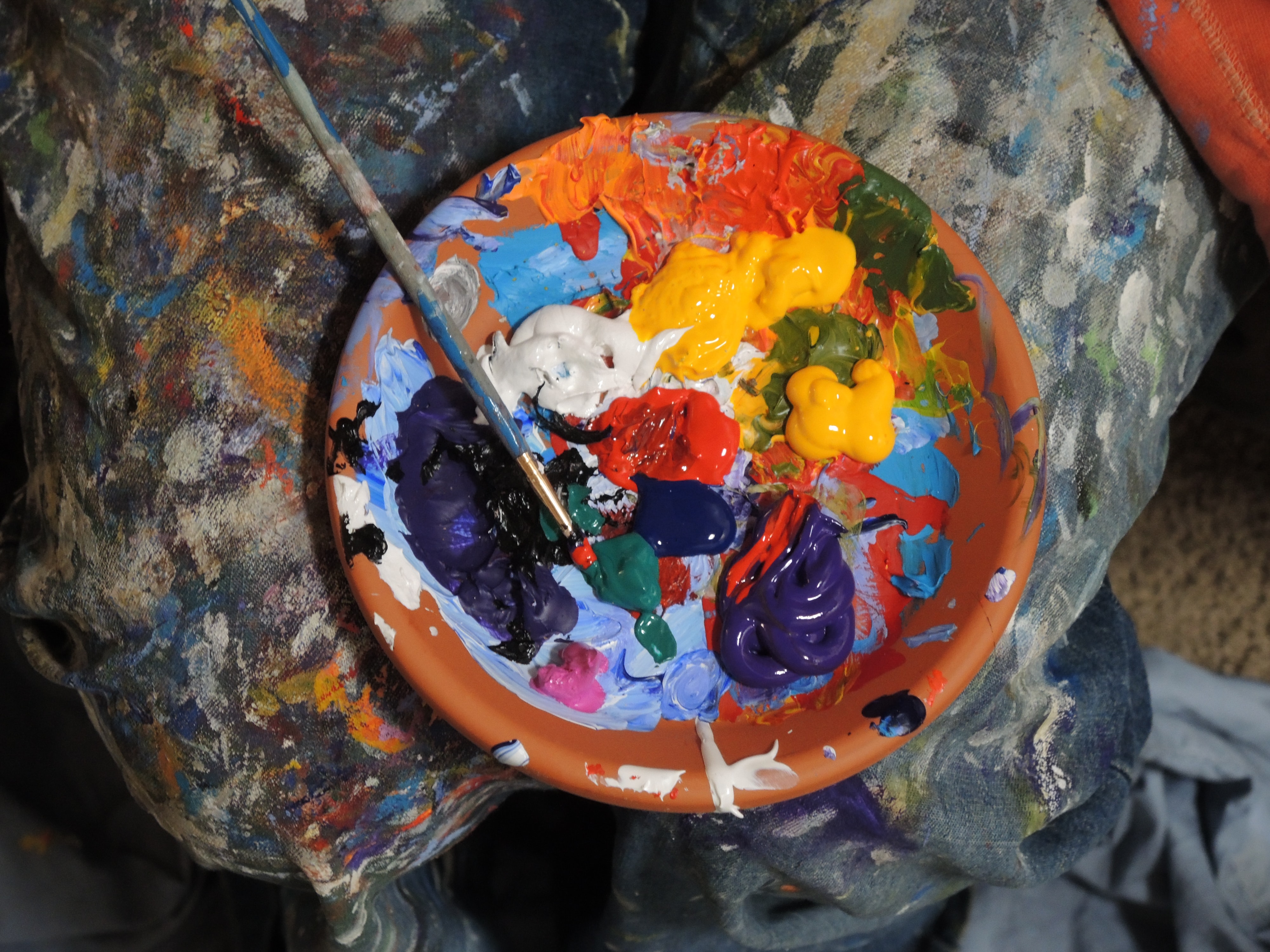 A bowl filled with different coloured paints, being used as a mixing palette. There's a paintbrush resting on the top and the bowl is sitting on a painted canvas sheet.