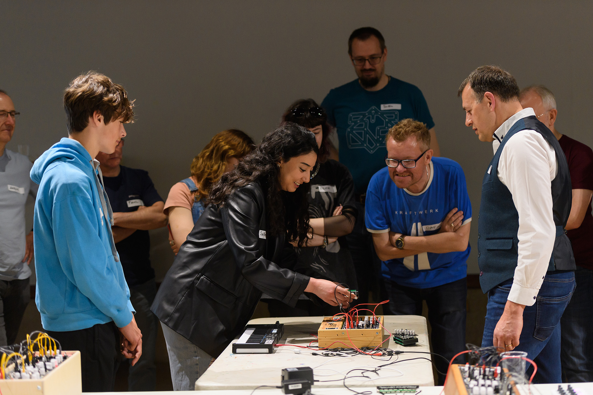 Rick Holt leading a modular synth workshop at the Herbert in 2022, with a group of people of all different ages gathered round watching a woman experimenting with a modular synth 